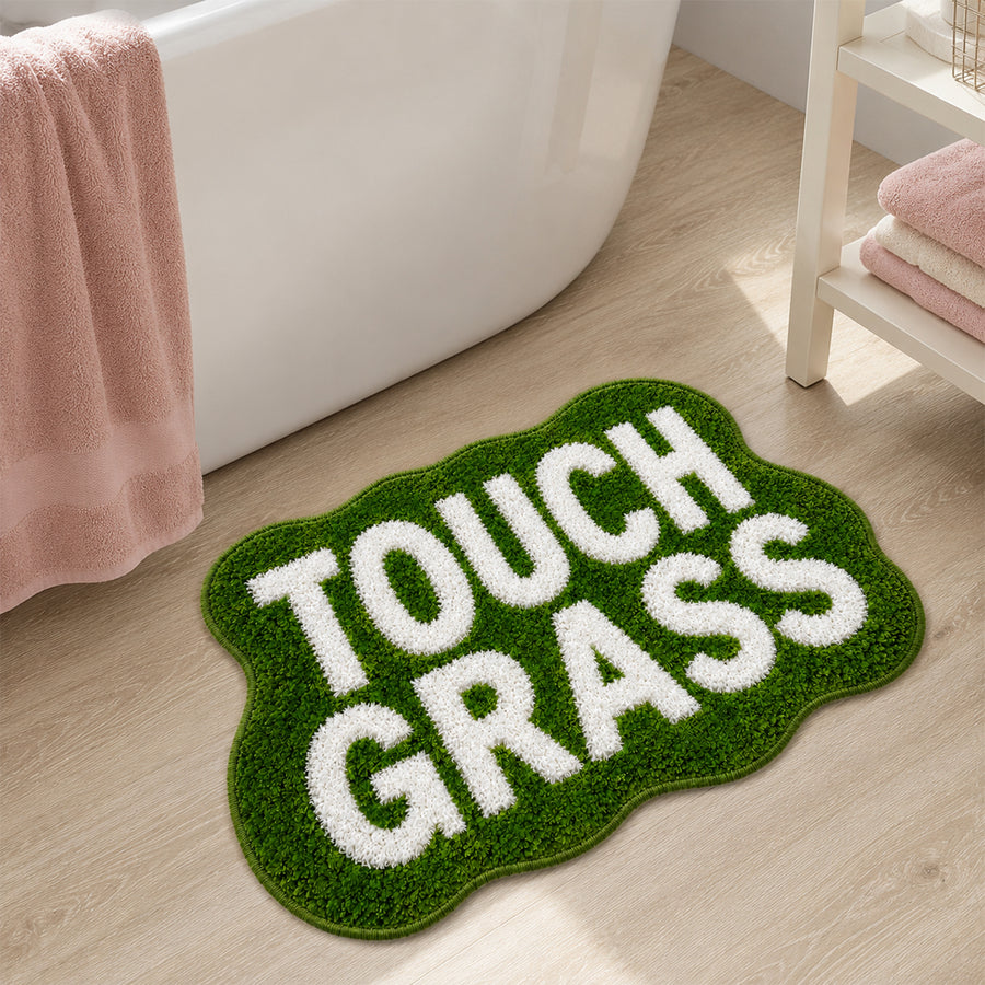 Green TOUCH GRASS bath mat on wood floor in front of a bathtub